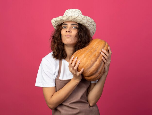 Foto gratuita jardinero de sexo femenino joven complacido en uniforme vistiendo sombrero de jardinería sosteniendo calabaza