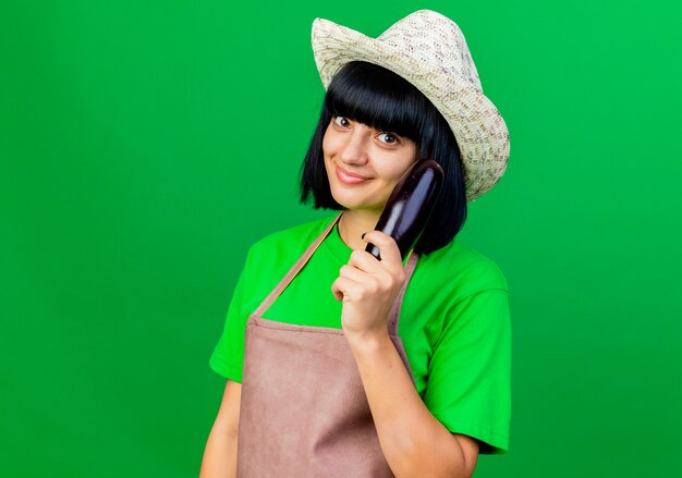 Jardinero de sexo femenino joven complacido en uniforme vistiendo sombrero de jardinería sostiene berenjena