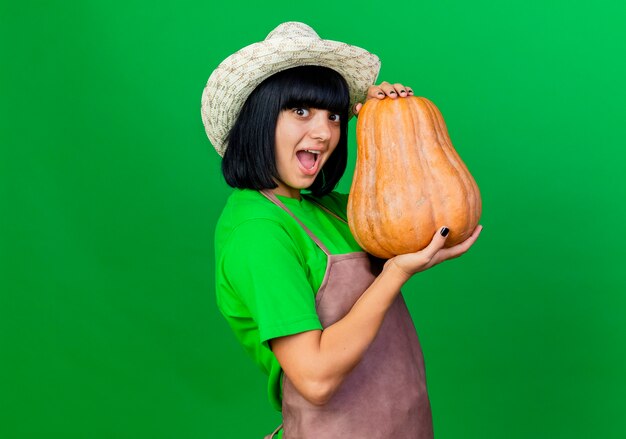 Jardinero de sexo femenino joven complacido en uniforme vistiendo sombrero de jardinería sostiene calabaza