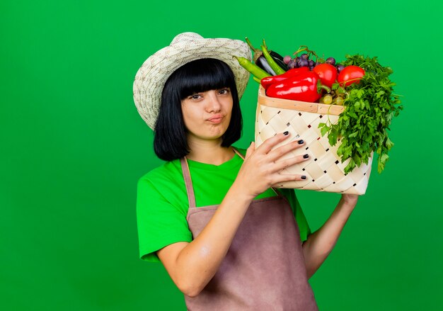 Jardinero de sexo femenino joven complacido en uniforme vistiendo sombrero de jardinería sostiene canasta de verduras