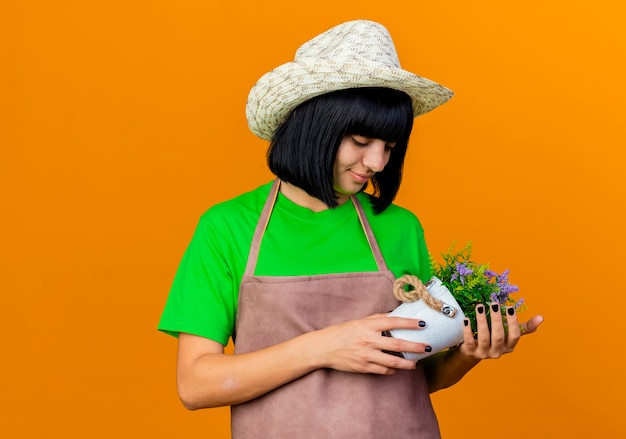 Jardinero de sexo femenino joven complacido en uniforme vistiendo sombrero de jardinería sostiene maceta