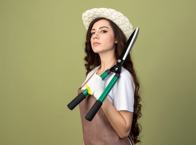 Jardinero de sexo femenino joven confiado en uniforme que lleva el sombrero y los guantes de jardinería se coloca de lado sosteniendo podadoras de jardín en el hombro aislado en la pared verde oliva