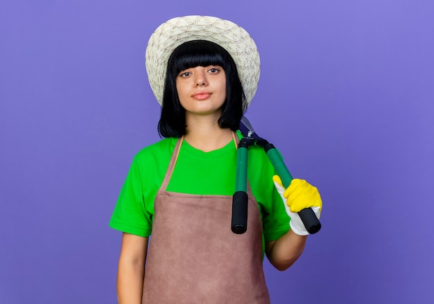 Jardinero de sexo femenino joven confiado en uniforme que lleva el sombrero y los guantes de jardinería sostiene las podadoras