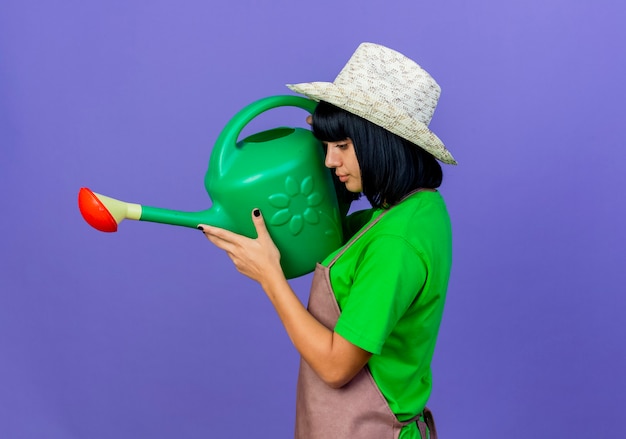 Jardinero de sexo femenino joven confiado en uniforme que lleva el sombrero de jardinería está de pie lateralmente sosteniendo la regadera