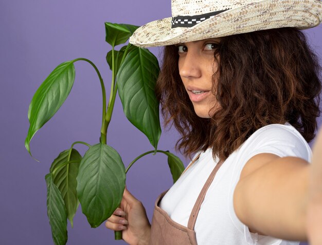 Jardinero de sexo femenino joven confiado en uniforme que lleva el sombrero de jardinería que sostiene la planta con la cámara aislada en púrpura