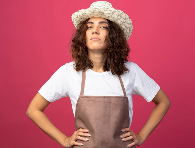 Foto gratuita jardinero de sexo femenino joven confiado en uniforme que lleva el sombrero que cultiva un huerto que pone las manos en la cadera aislada en rosa