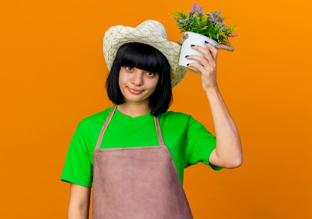 Foto gratuita jardinero de sexo femenino joven confiado en uniforme que lleva el sombrero que cultiva un huerto sostiene la maceta