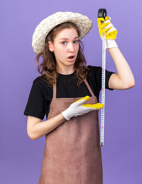 Jardinero de sexo femenino joven confundido con sombrero de jardinería con guantes de estiramiento y puntos en la cinta métrica