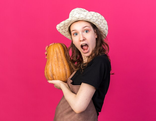 Jardinero de sexo femenino joven emocionado con sombrero de jardinería sosteniendo calabaza aislado en la pared rosa