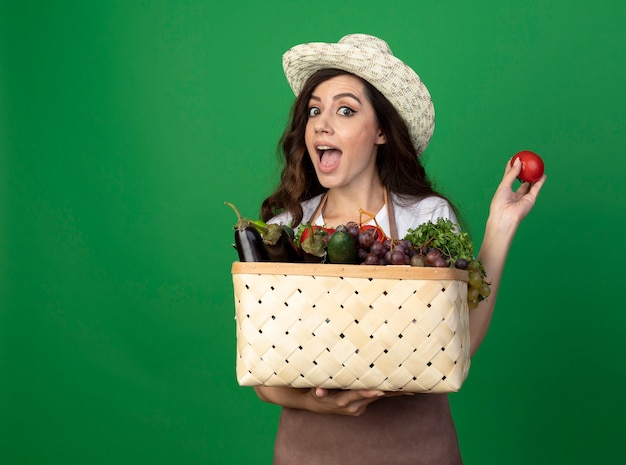 Jardinero de sexo femenino joven emocionado en uniforme con sombrero de jardinería sostiene cesta de verduras y tomate aislado en la pared verde