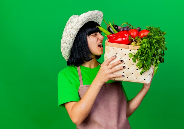 Jardinero de sexo femenino joven enojado en uniforme que lleva el sombrero de jardinería sostiene y mira canasta de verduras