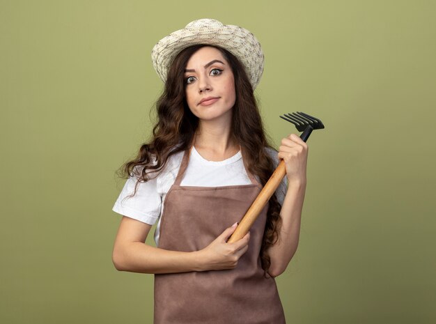 Jardinero de sexo femenino joven impresionado en uniforme que lleva el sombrero de jardinería sostiene el rastrillo y mira al frente aislado en la pared verde oliva