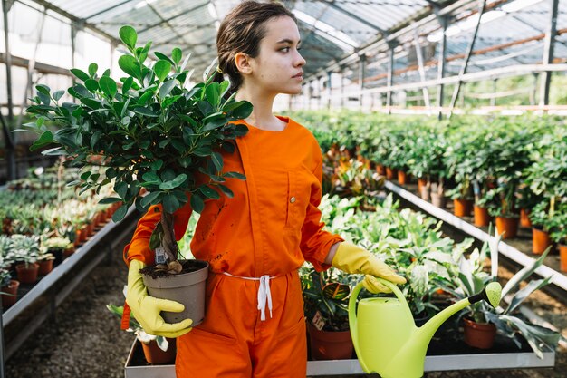 Jardinero de sexo femenino joven que sostiene la planta y la regadera en conserva en invernadero