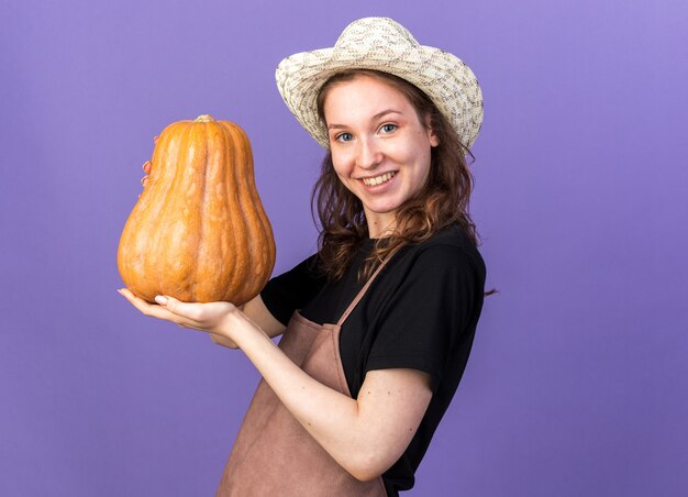 Jardinero de sexo femenino joven sonriente que lleva el sombrero de jardinería que sostiene la calabaza aislada en la pared azul