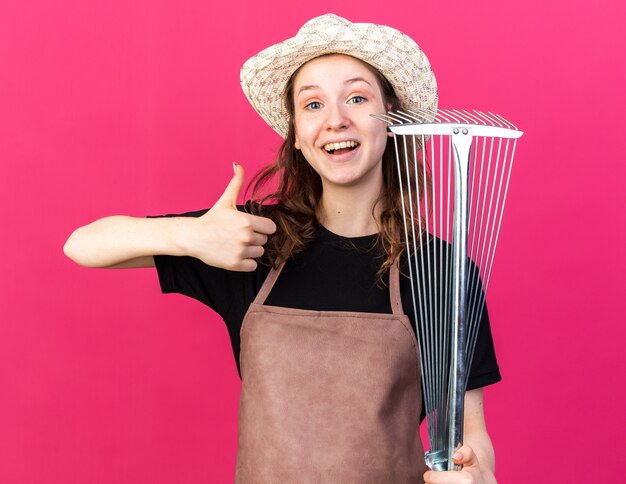 Jardinero de sexo femenino joven sonriente que lleva el sombrero de jardinería que sostiene el rastrillo de la hoja que muestra el pulgar hacia arriba aislado en la pared rosada