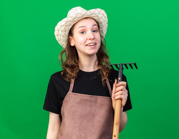 Jardinero de sexo femenino joven sonriente que lleva el sombrero que cultiva un huerto que sostiene el rastrillo