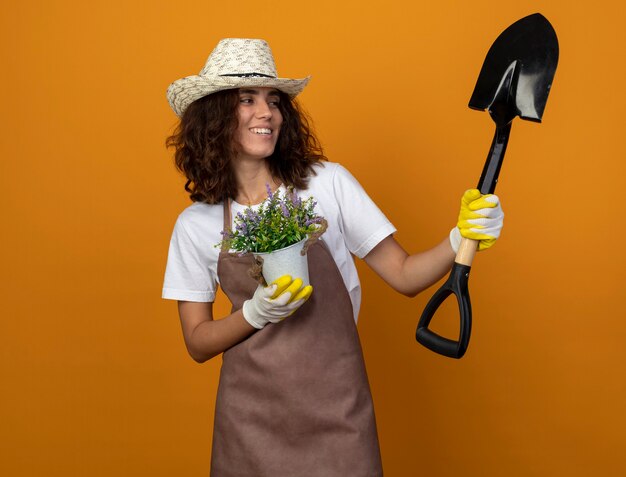 Jardinero de sexo femenino joven sonriente en uniforme que lleva el sombrero y los guantes de jardinería que sostienen la flor en la maceta y que mira la pala en su mano