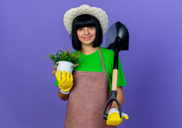 Jardinero de sexo femenino joven sonriente en uniforme que lleva el sombrero y los guantes de jardinería sostiene la pala