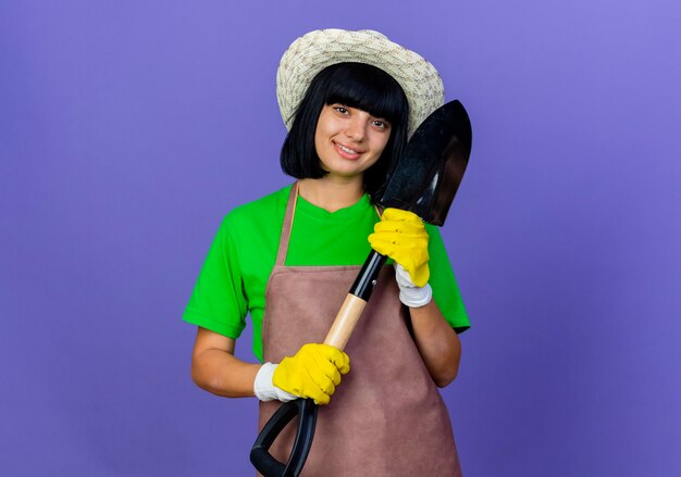 Jardinero de sexo femenino joven sonriente en uniforme que lleva el sombrero y los guantes de jardinería sostiene la pala