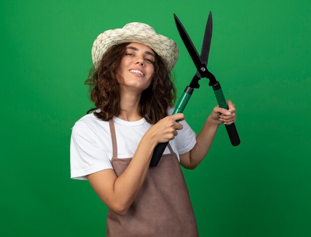 Jardinero de sexo femenino joven sonriente en uniforme que lleva el sombrero de jardinería levantando tijeras