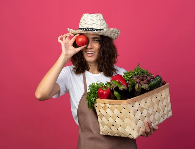 Jardinero de sexo femenino joven sonriente en uniforme que lleva el sombrero de jardinería que sostiene la cesta de verduras que muestra el gesto de la mirada con el tomate aislado en rosa