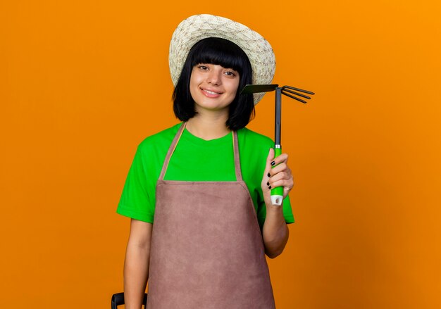 Jardinero de sexo femenino joven sonriente en uniforme que lleva el sombrero de jardinería sostiene la pala y el rastrillo de la azada