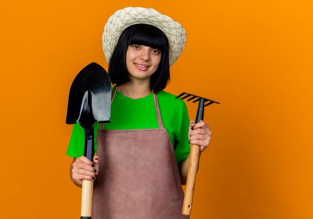 Jardinero de sexo femenino joven sonriente en uniforme que lleva el sombrero de jardinería sostiene la pala y el rastrillo