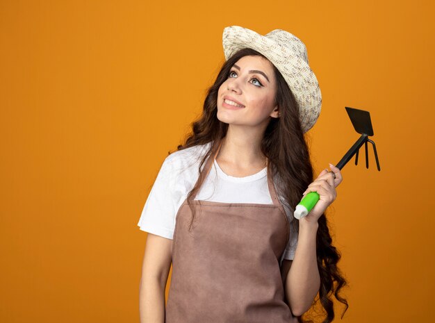 Jardinero de sexo femenino joven sonriente en uniforme que lleva el sombrero de jardinería sostiene el rastrillo de la azada y mira el lado aislado en la pared naranja con espacio de copia