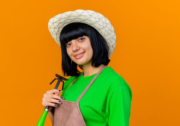 Foto gratuita jardinero de sexo femenino joven sonriente en uniforme que lleva el sombrero que cultiva un huerto que sostiene el rastrillo de la azada