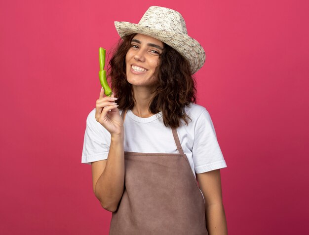 Jardinero de sexo femenino joven sonriente en uniforme que lleva el sombrero que cultiva un huerto sosteniendo la pimienta que rompe aislada en rosa