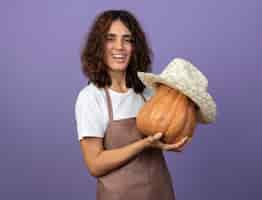 Foto gratuita jardinero de sexo femenino joven sonriente en uniforme que sostiene la calabaza con el sombrero