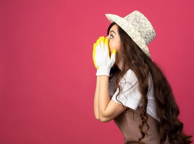 Jardinero de sexo femenino joven sorprendido en uniforme que lleva el sombrero y los guantes de jardinería se coloca de lado poniendo las manos en la boca aislada en la pared rosada con el espacio de la copia