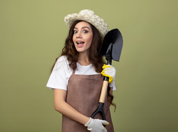 Jardinero de sexo femenino joven sorprendido en uniforme que lleva el sombrero y los guantes de jardinería sostiene la pala aislada en la pared verde oliva