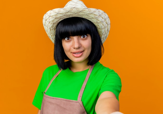 Foto gratuita jardinero de sexo femenino joven sorprendido en uniforme que lleva el sombrero de jardinería pretende sostener la cámara