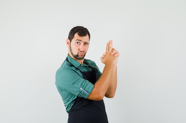 Jardinero de sexo masculino en camiseta, delantal que muestra el gesto de la pistola de tiro y parece seguro, vista frontal.