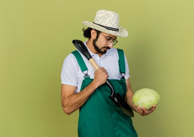 Jardinero de sexo masculino confiado en gafas ópticas con sombrero de jardinería sostiene la pala y mira el repollo