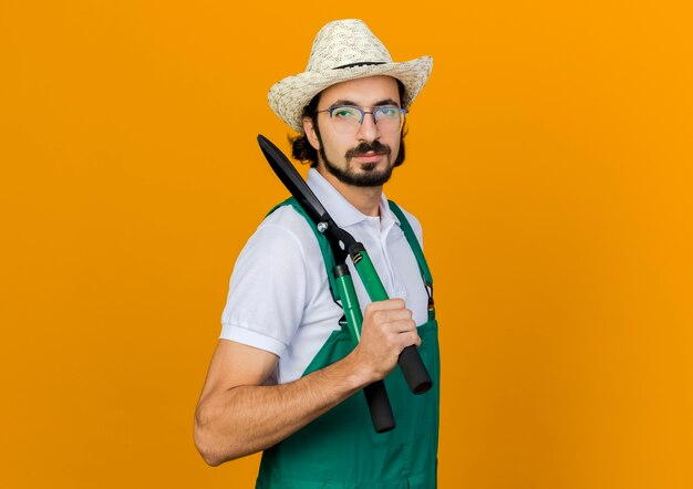 Jardinero de sexo masculino confiado en gafas ópticas con sombrero de jardinería sostiene tijeras en el hombro