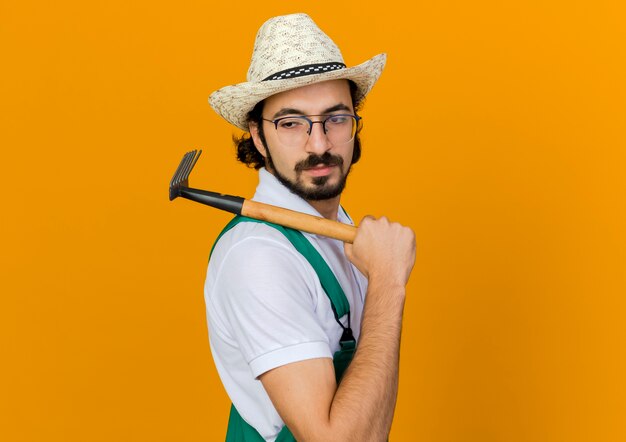 Jardinero de sexo masculino confiado en gafas ópticas con sombrero de jardinería tiene rastrillo de azada en el hombro