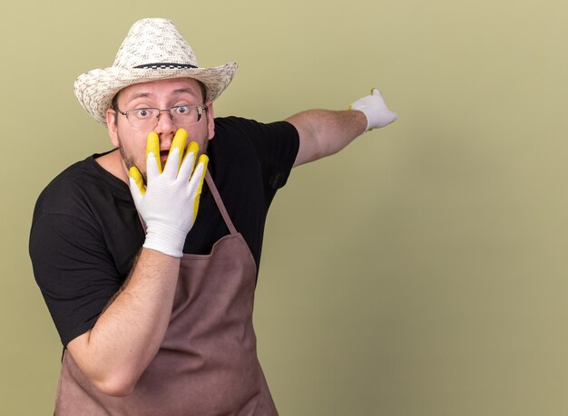 Jardinero de sexo masculino joven asustado con puntos de guantes y sombrero de jardinería detrás de la boca cubierta con la mano aislada en la pared verde oliva con espacio de copia