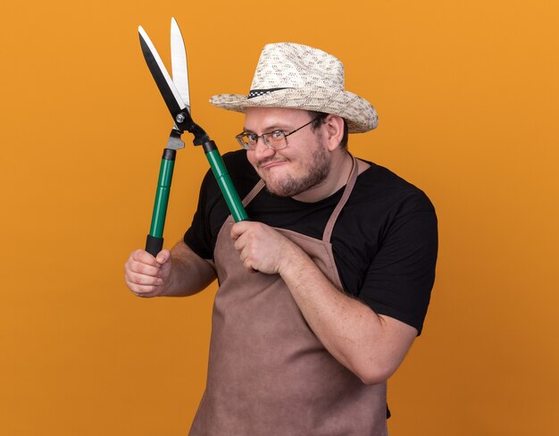 Jardinero de sexo masculino joven codicioso con sombrero y guantes de jardinería sosteniendo tijeras aislado en la pared naranja