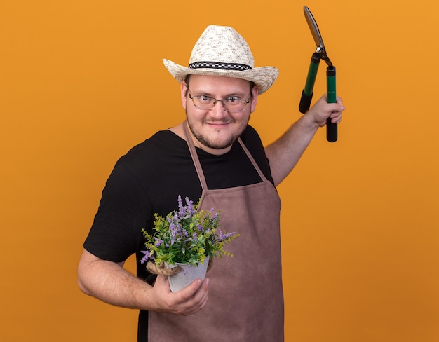 Jardinero de sexo masculino joven complacido con sombrero y guantes de jardinería sosteniendo tijeras con flor en maceta extendiendo la mano aislada en la pared naranja