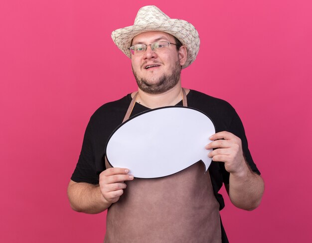 Jardinero de sexo masculino joven complacido con sombrero de jardinería sosteniendo bocadillo aislado en la pared rosa