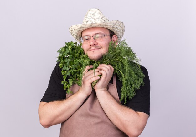 Jardinero de sexo masculino joven complacido con sombrero de jardinería sosteniendo eneldo con cilantro alrededor de la cara aislada en la pared blanca
