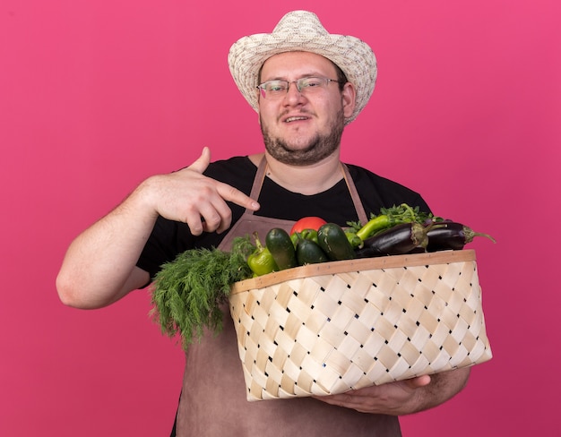 Jardinero de sexo masculino joven complacido con sombrero de jardinería sosteniendo y puntos en la canasta de verduras aislado en la pared rosa