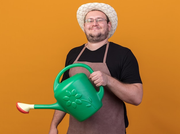 Jardinero de sexo masculino joven complacido con sombrero de jardinería sosteniendo regadera aislado en la pared naranja