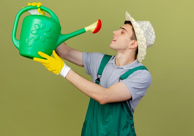 Jardinero de sexo masculino joven complacido vistiendo guantes y gorro de jardinería tiene regadera