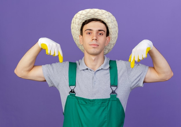 Jardinero de sexo masculino joven confiado con guantes y sombrero de jardinería apunta hacia abajo