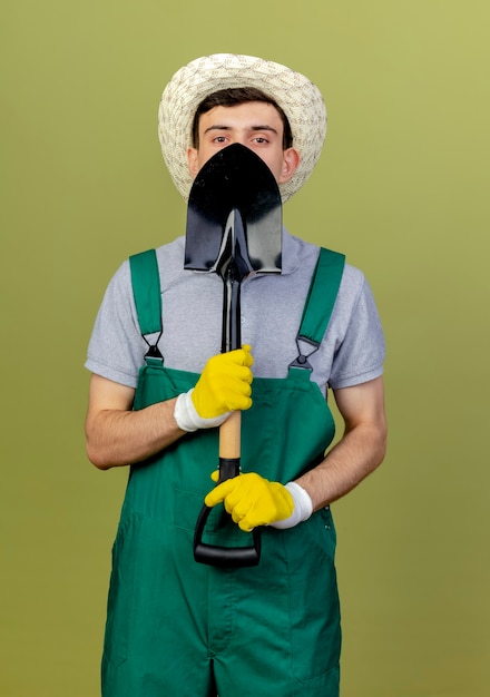 Foto gratuita jardinero de sexo masculino joven confiado que lleva el sombrero y los guantes de jardinería sostiene la pala
