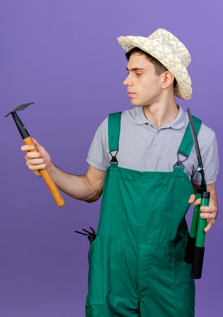 Jardinero de sexo masculino joven confiado con sombrero de jardinería se encuentra con herramientas de jardinería mirando rastrillo aislado sobre fondo púrpura con espacio de copia