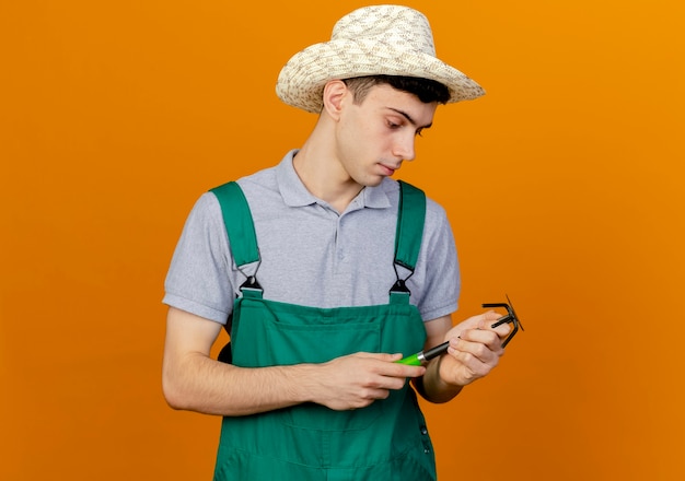 Jardinero de sexo masculino joven confiado con sombrero de jardinería mira rastrillo de azada